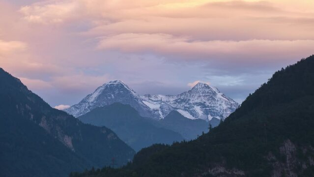 Switzerland snow capped mountain peaks at sunset, with layers of lenticular clouds forming above and warm pink color cast over hills, in timelapse. Swiss nature, landscape.