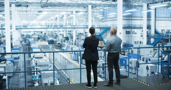 Portrait of a Middle Aged Engineer and Happy Female Scientist Using Laptop Computer and Talking in a Factory Facility with Equipment Producing Modern Technical Devices for Different Industries