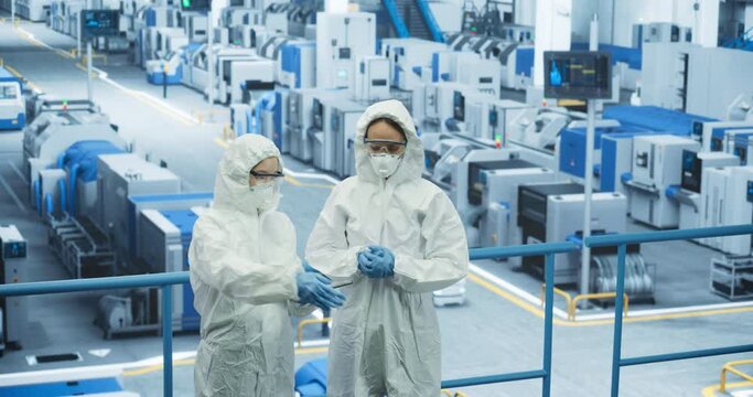 Female Workers Wearing Full-Body Polypropylene Coveralls at a Manufacturing Plant. Specialists Using Tablet Computer, Discussing Automated Work Process. Employees Protected From Hazardous Materials