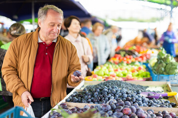 Positive mature male holding plum while choosing fruits and vegetables at market