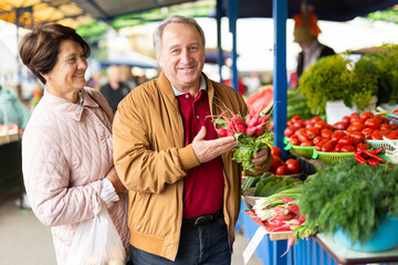 Positive mature couple in casual wear picking fresh radish during shopping at crowded bazaar