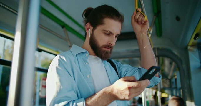 Young man using smartphone, listening dance energetic music, get messages while riding in modern tram. Attractive guy wearing wireless headphones, holding handrail, enjoying bus trip at sunny day.