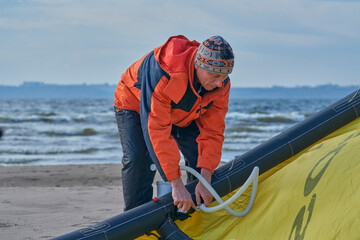 A mature male surfer is preparing to ride a bosque with a kite on the sandy shore of a lake. Cold windy autumn day.