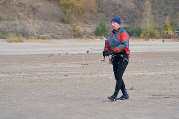 A mature male surfer is preparing to ride a bosque with a kite on the sandy shore of a lake. Cold windy autumn day.