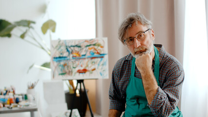 Fototapeta premium Man Artist In An Apron Sits Thoughtfully On Chair In Studio And Looks At Camera
