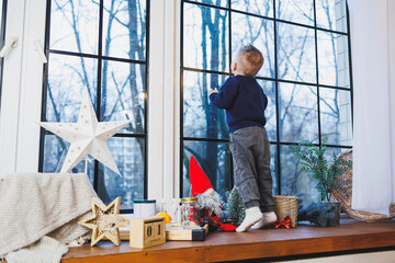 A 2-year-old boy sits on the windowsill in the new year. New Year's atmosphere at home. The child is waiting for the new year.