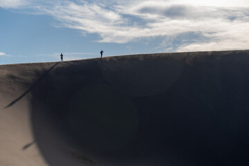 People hike up High Dune at Great Sand Dunes National Park in Colorado on a sunny summer evening, with mountains in the background - obrazy, fototapety, plakaty