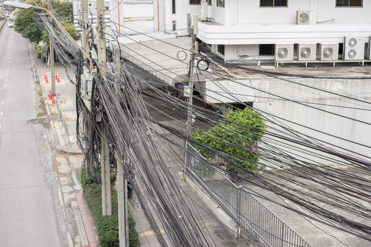 On the side of a road in Thailand there is an electric pole with a mess of wires and buildings and trees behind it.
