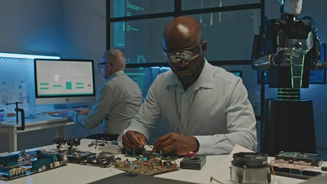 Medium shot of African American engineer soldering parts of human robot while working at desk in modern futuristic lab at night with colleague