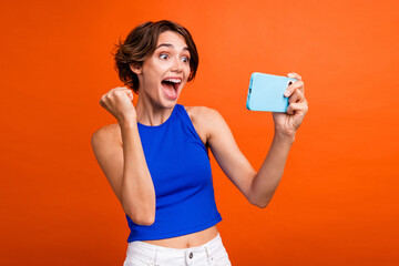 Portrait of delighted pretty lady raise fist hold smart phone playing games eshopping isolated on orange color background