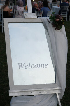 A white board with the inscription welcome stands on a green lawn. Signboard with the invitation of guests to the wedding ceremony and banquet. Beautiful wedding decor decorated with flowers