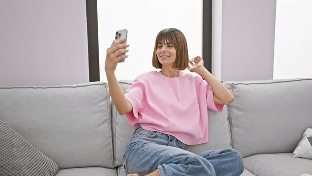 Joyful young hispanic woman enjoys taking a beautiful selfie sitting comfortably on her sofa at home, her happiness and smile shining through the photo