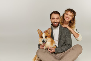 family portrait, happy man and woman in winter attire posing with cute corgi on grey background