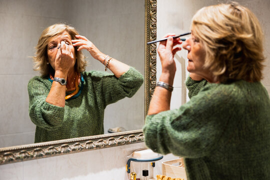 Older Woman Applying Eye Make-up In The Bathroom Mirror