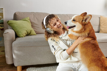 curly woman in wireless headphones playing with cute corgi dog in modern apartment, happy moments - obrazy, fototapety, plakaty