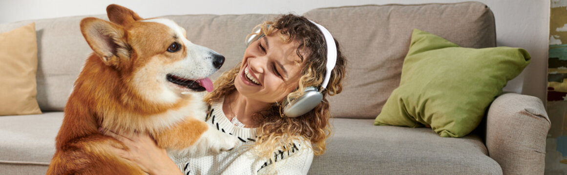 Curly Woman In Wireless Headphones Playing With Cute Corgi Dog In Modern Apartment, Happy Moments