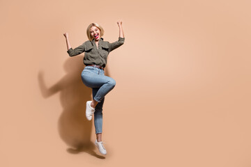 Fototapeta na wymiar Full length photo of excited lucky lady wear khaki shirt jumping high rising fists empty space isolated beige color background