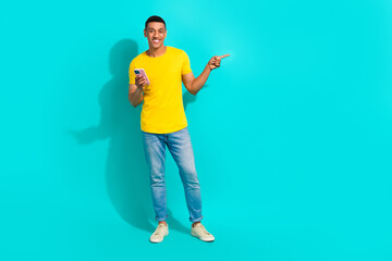 Fototapeta na wymiar Full length portrait of cheerful handsome guy hold smart phone direct finger empty space isolated on turquoise color background