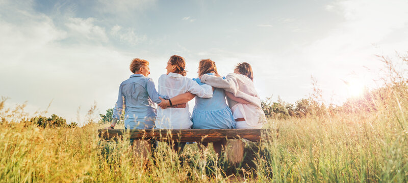 ..Four smiling women dressed light summer clothes harm embracing sitting on the meadow bench during outdoor walking. Woman's friendship, relations, and happiness concept image.