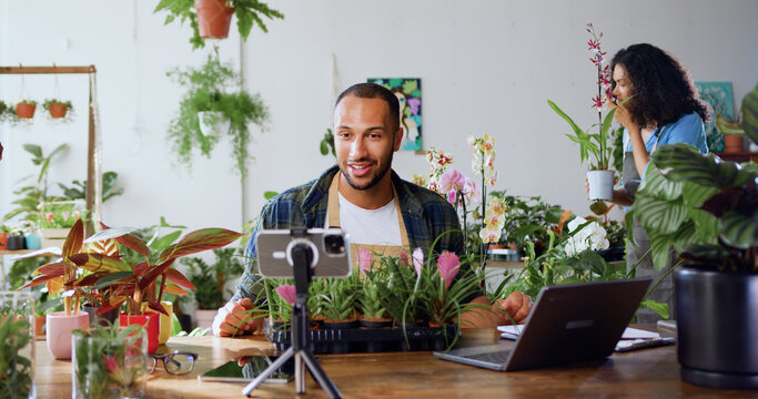 Handsome man gardener in apron using phone and recording blog video with clients. Young adult content creator influencer blogger recording a video talking about indoor plants care from workplace.