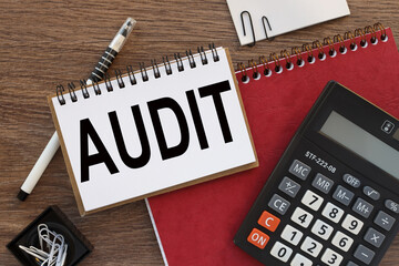 Audit. A blank red notebook with a pen lies on a wooden office table near a calculator and a white...