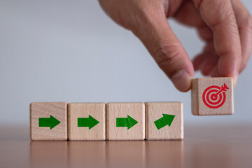 Arrow leading point to target icon on wooden blocks with businessman hand placing red goal dartboard symbol. Business agenda and mission setting concept. Dart board cube future vision strategy success