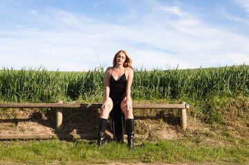Blonde woman with long hair, young and beautiful dressed in black dress and black boots sitting sensually on wooden fence. In the background a green wheat field and blue sky with white clouds.