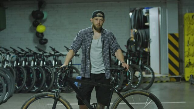 Medium long shot of young man standing with bicycle in garage and looking at camera
