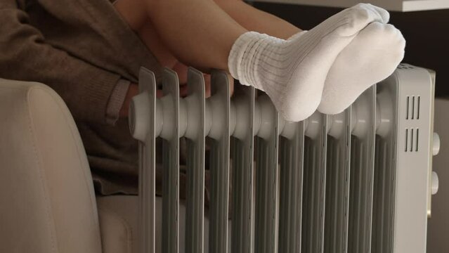 Woman in warm clothes is sitting next to the radiator. Woman warming her feet on an electric heater. It's cold at home. Energy crisis and high heating prices.