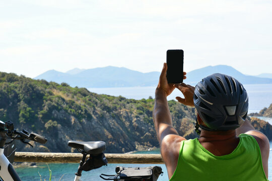 A middle-aged man in a green T-shirt and helmet on an electric bicycle stops and takes pictures with a black phone of the beautiful Italian landscape on the Tuscan coast during his bike tour.