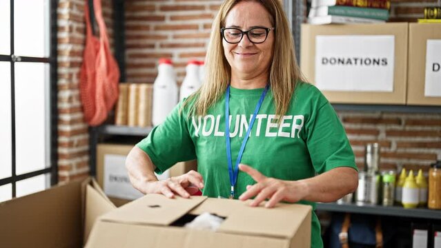 Middle age hispanic woman volunteer smiling confident packing clothes on cardboard box to donate at charity center