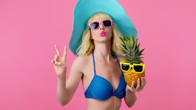 Sexy woman in bikini, peace hand, dancing with pineapple on pink, fashionable hat, smiling enjoys summertime for spa, wellness. Playful slim lady dance for rest, tan or pool party, colorful trend
