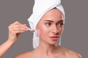 Young woman massaging her face with rose quartz roller on grey background