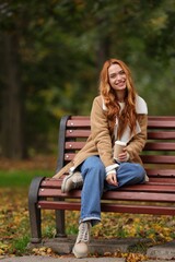 Smiling woman with paper cup sitting on bench in autumn park