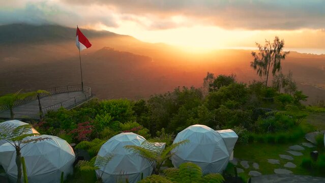 A girl stands by the infinity pool during sunsrise in Glamping camping against the backdrop of the Batur volcano in Kintamani, Bali, Indonesia. Aerial drone view.