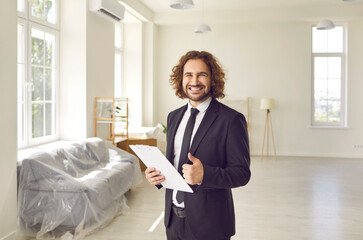 Portrait of friendly young male realtor showing thumbs up recommending his real estate agency. Young millennial man in suit stands in apartment with clipboard and smiles at camera with big smile. - obrazy, fototapety, plakaty