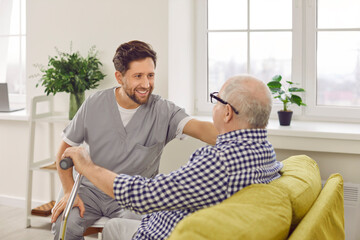 Optimistic male caregiver comforting senior patient, touching his shoulder. Male carer in uniform giving support to elderly man who sitting on sofa with walking cane. Elderly healthcare, medical care - obrazy, fototapety, plakaty