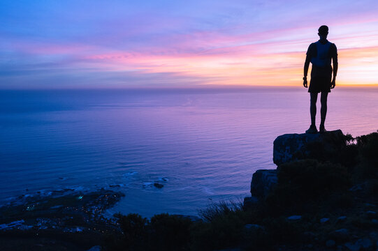 A hiker silhouetted against the sunset over the ocean while standing on the top of a mountain that they have successfully climbed.
