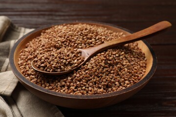 Bowl and spoon with dry buckwheat on wooden table, closeup