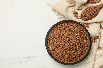 Bowl with dry buckwheat, spoon and fabric on white marble table, flat lay. Space for text
