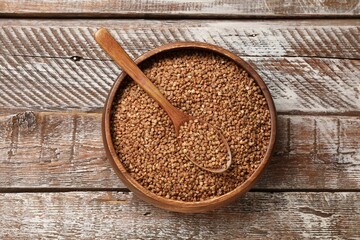 Bowl and spoon with dry buckwheat on wooden table, top view
