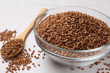 Bowl and spoon with dry buckwheat on white tiled table, closeup