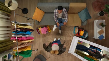Top view of room. Man on the sofa, girl posing in new leather jacket and hat in front of the mirror, man confused, girl doesn't like outfit.