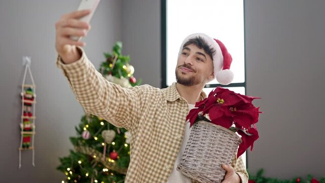 Young arab man make selfie by smartphone holding plant standing by christmas tree at home