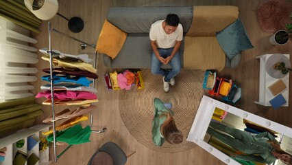 Top view of apartment living room. Man sits on the sofa, happy girl posing in new pajamas in front of the mirror, man likes the outfit.