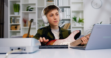 Attractive 13-14 years boy wearing headphones playing guitar looking at the laptop at home. Portrait of teenage learning to play e guitar by watching video tutorial on laptop - obrazy, fototapety, plakaty