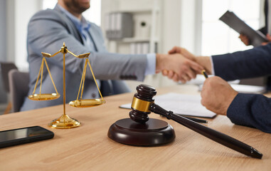 Closeup close up shot of judge's gavel, justice scales and mobile phone on office table, with lawyer attorney and client exchanging handshakes in background. Legal services, law consultation concept