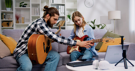 Music teacher and teenage student girl sitting on sofa learning to play new song on guitar in...