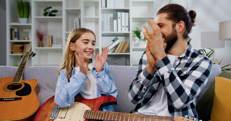 Handsome man sitting on the sofa and teaching his small daughter playing the guitar, girl taking...
