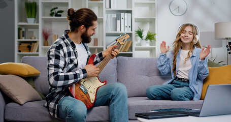 Happy Father's day. Portrait of smiling father playing guitar while has daughter in headphones dancing and sitting sofa at home, Activity family love and lifestyle concept - obrazy, fototapety, plakaty
